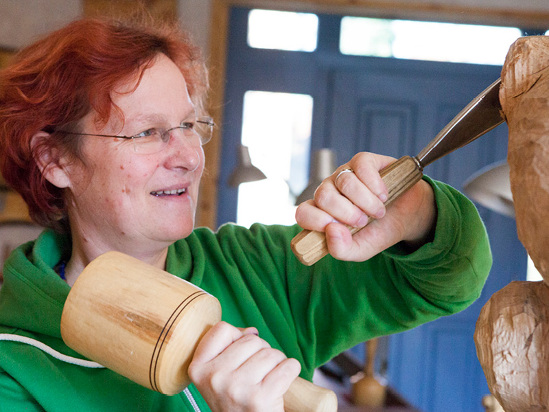 Felice Meer arbeitet als Holzbildhauerin mit Holzhammer und Schnitzeisen an einer Holzskulptur in der Werkstatt
